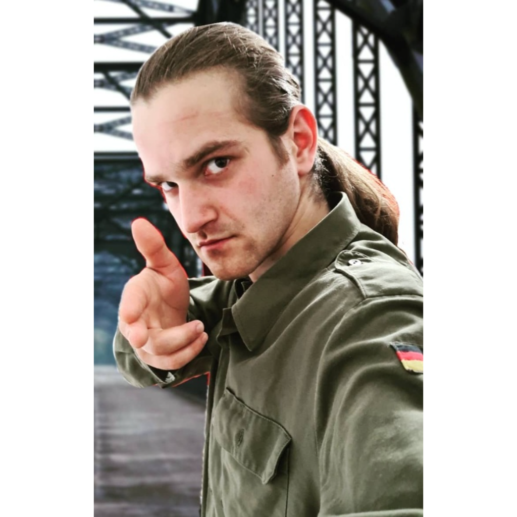 A young man with light brown hair in a ponytail, wearing a green military-style shirt with a German flag patch, points his finger towards the camera against a black metal structure.